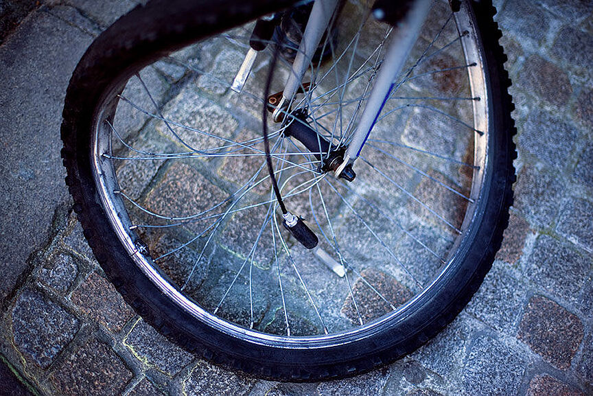 Verbogene Fahrradfelge Nahaufnahme eines Fahrradvorderrads auf Kopfsteinpflaster. Die Felge ist verbogen und mehrere Speichen sind gebrochen oder hängen lose herab. Das Rad steht schief auf dem grauen Pflaster und ist offensichtlich nach einem Unfall nicht mehr fahrbereit.